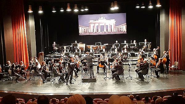 Blick auf die Bühne mit den Symphonikern und Dirigent; Foto vom Brandenburger Tor im Hintergrund
